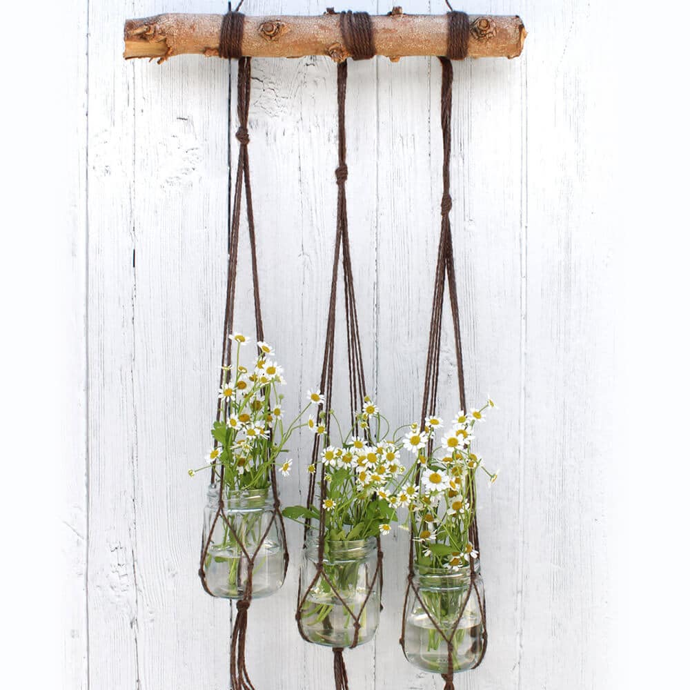 Fotografía estilizada de tres macetas llenas de flores que cuelgan de tres colgadores de macramé para plantas